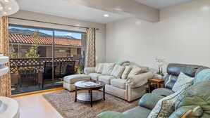 Una sala de estar moderna con un sofá, un sillón y una mesita. Se ve un balcón con barandilla y un edificio con tejado de tejas.