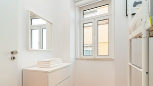 Een kleine, witte kamer met een houten vloer, een wit kastje met opgevouwen handdoeken en een raam met luiken.