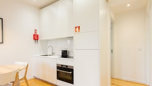 A compact kitchen with white cabinets, a sink, a stove, and a refrigerator. There is a small table with chairs and a fire extinguisher mounted on the wall. - Arroios Concept IV by Homing (Lisbon)