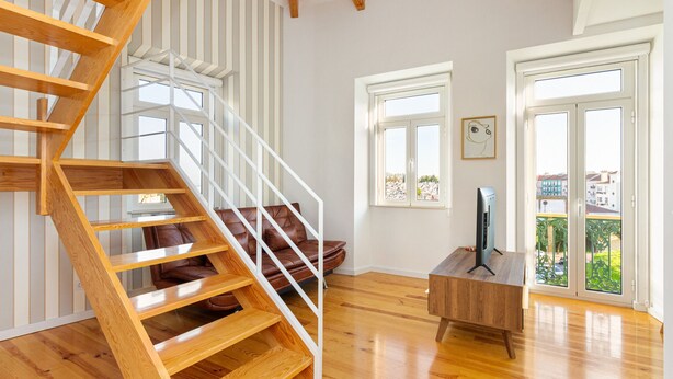 Une pièce spacieuse avec un escalier en bois, un canapé en cuir, une table basse en bois et une grande fenêtre donnant sur des immeubles.