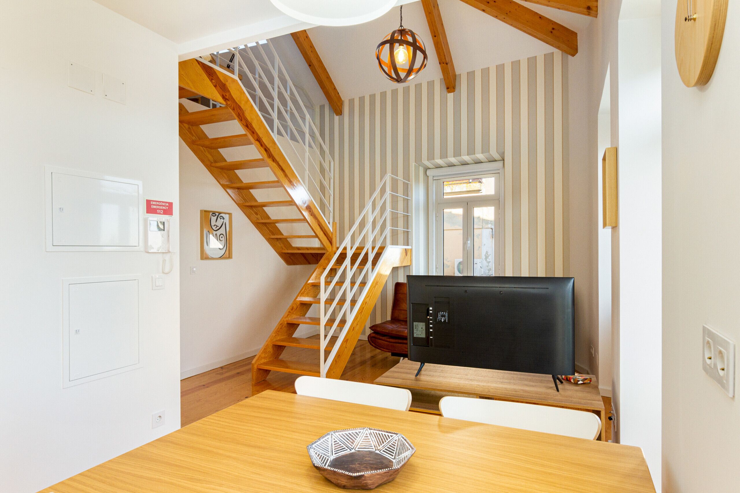 A dining area with a wooden table, chairs, and a decorative bowl. A staircase with wooden railings leads to an upper level.