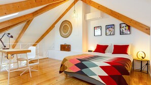 Une chambre avec un plafond en pente, des poutres en bois, un lit recouvert d’une courtepointe colorée, un bureau avec une chaise, une lampe fixée au mur et des œuvres d’art.