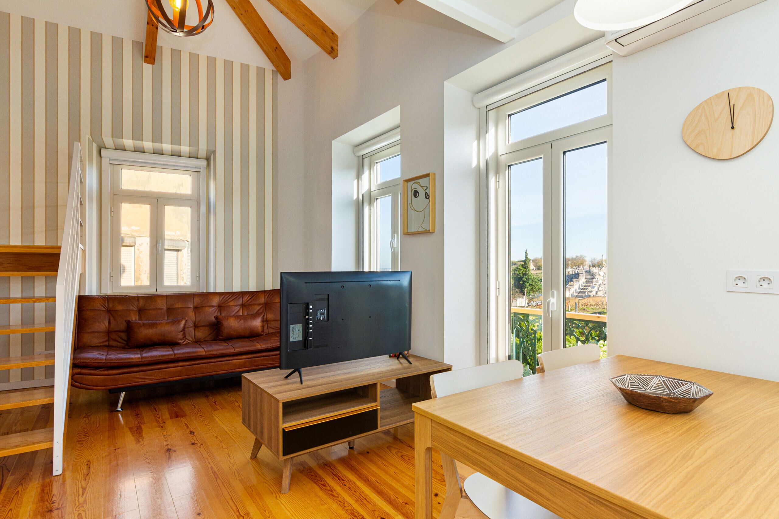 Una sala de estar con piso de madera, un sofá de cuero marrón, un televisor sobre un mueble de madera y una mesa de comedor con un tazón.