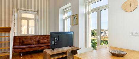 Una sala de estar con piso de madera, un sofá de cuero marrón, un televisor sobre un mueble de madera y una mesa de comedor con un tazón.