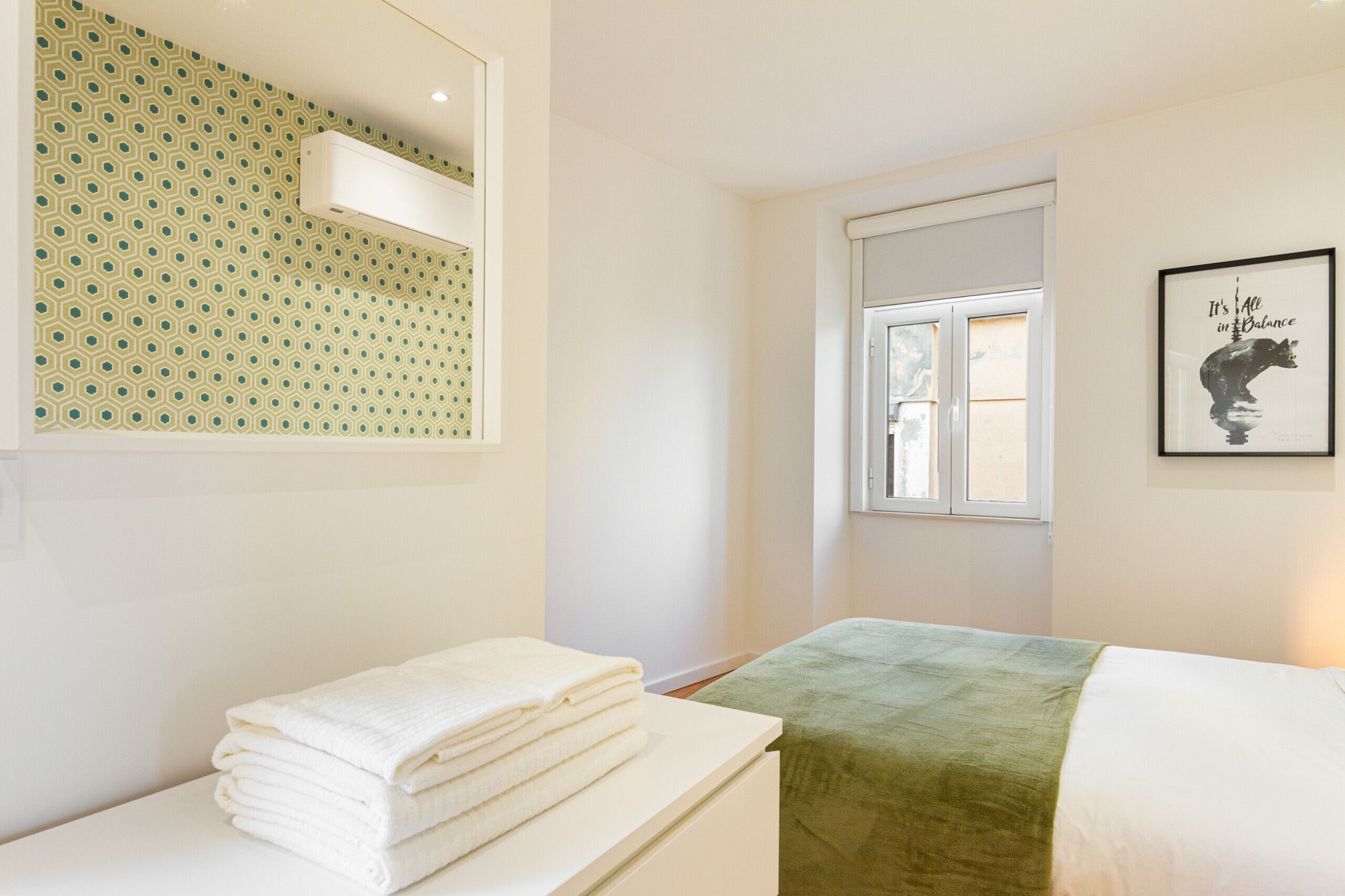 A hotel room with a bed, a window, a mirror, a towel stack, and a framed picture on the wall.