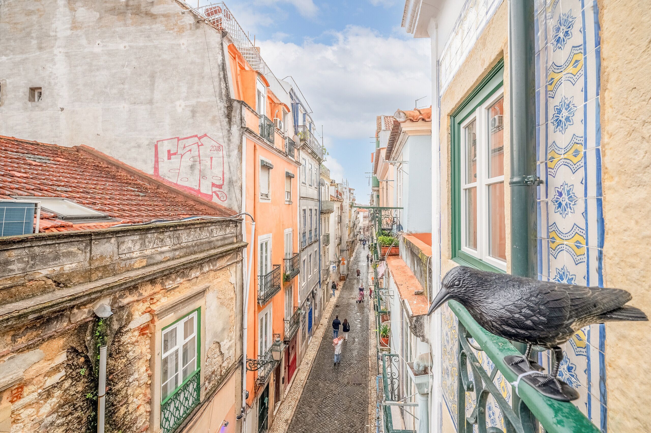 A narrow street with buildings on either side, a pigeon on a railing, and people walking.