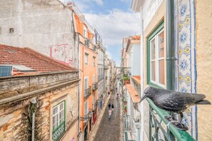 Uma rua estreita com edifícios em ambos os lados, uma pomba em um corrimão e pessoas caminhando.