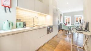 A modern kitchen with white cabinets, a stainless steel sink, and a built-in oven. There is a dining area with a table and chairs, and a window with curtains. - Bairro Alto Prime Duplex by Homing (Lisbon)