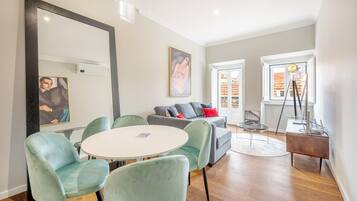 Un salon moderne avec une table ronde, deux fauteuils verts, un canapé gris avec des coussins rouges, un grand miroir et un tableau accroché au mur.