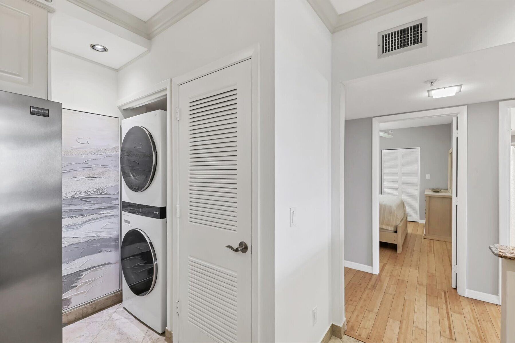 A laundry room with a Frigidaire washer and dryer, a white door with a louvered design, and a hallway leading to a bedroom with a bed and a nightstand.