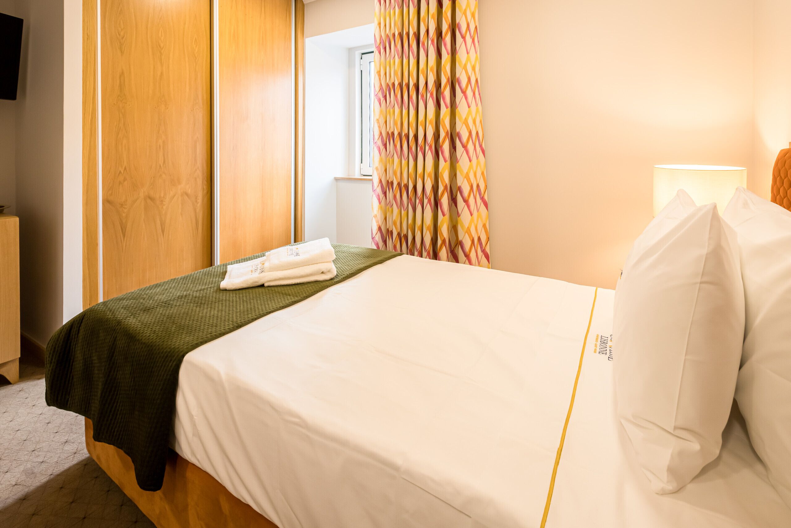 Une chambre d’hôtel avec un lit, une couverture verte, des oreillers blancs, une lampe de chevet et une fenêtre avec des rideaux.