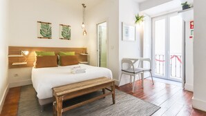 Ein gut beleuchtetes Schlafzimmer mit einem Bett, einem Nachttisch und einer Bank. Ein Sessel steht in der Nähe einer Schiebetür, die auf einen Balkon führt.