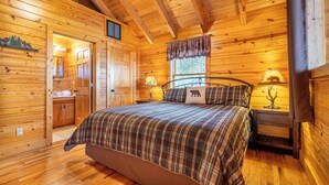 Ein gemütliches Schlafzimmer in einer Holzhütte mit einem großen Bett, zwei Nachttischen mit Lampen, einem Fenster mit Jalousien und einer Holztür.