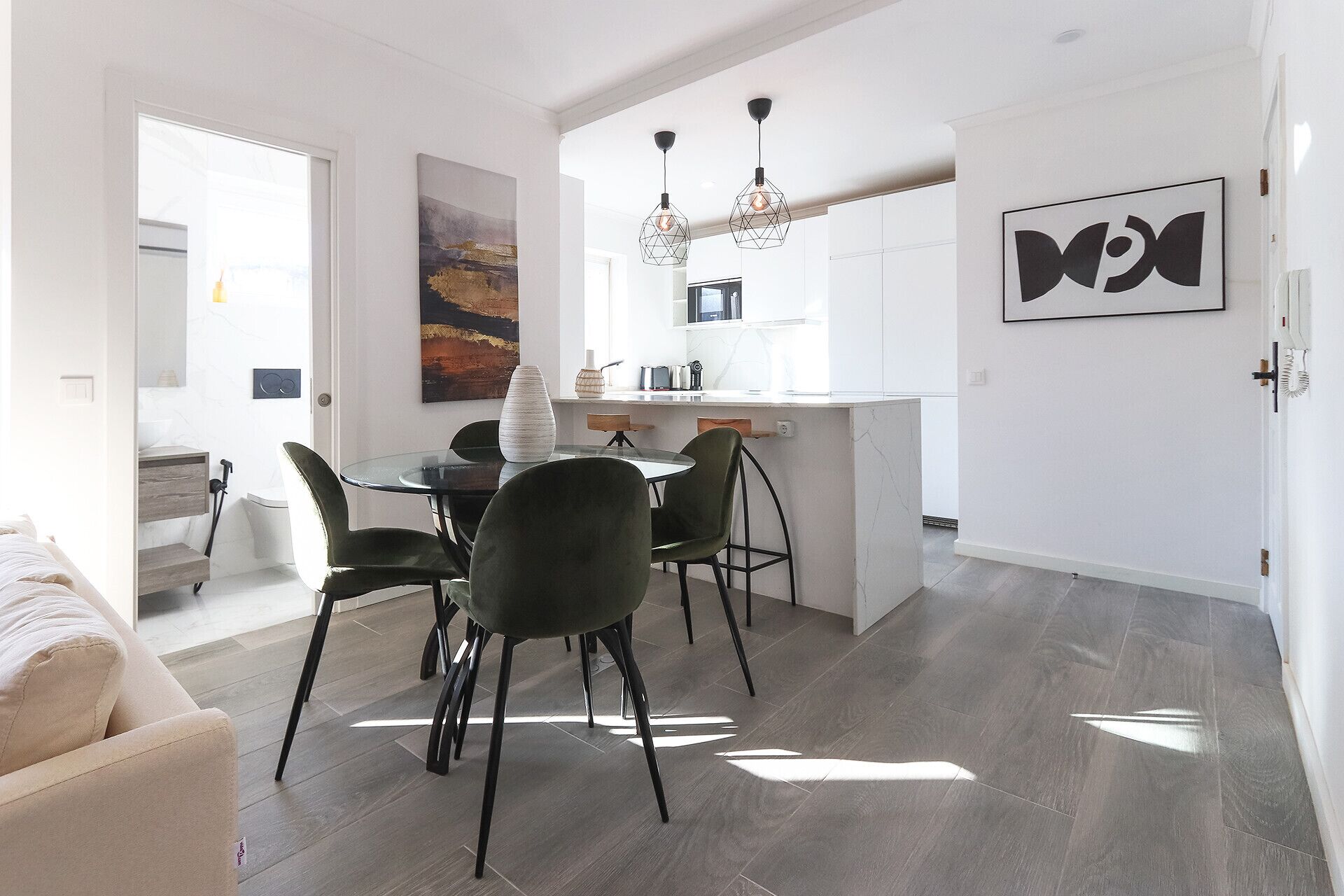 Una cocina moderna con armarios blancos, un comedor con mesa de vidrio y sillas negras, y una sala de estar con sofá y un cuadro en la pared.