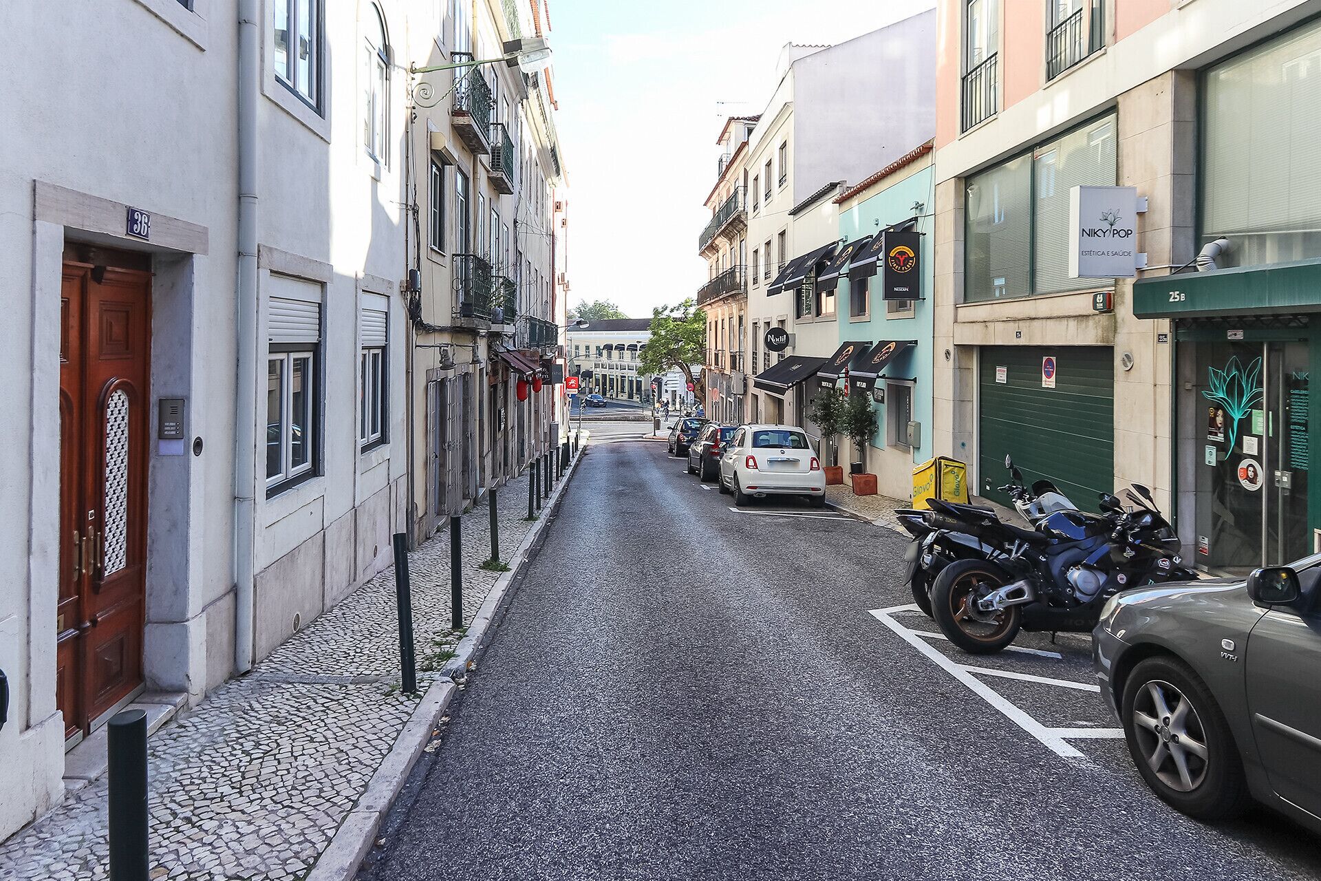 Eine enge Kopfsteinpflasterstraße gesäumt von Gebäuden, parkenden Autos und einigen Motorrädern.
