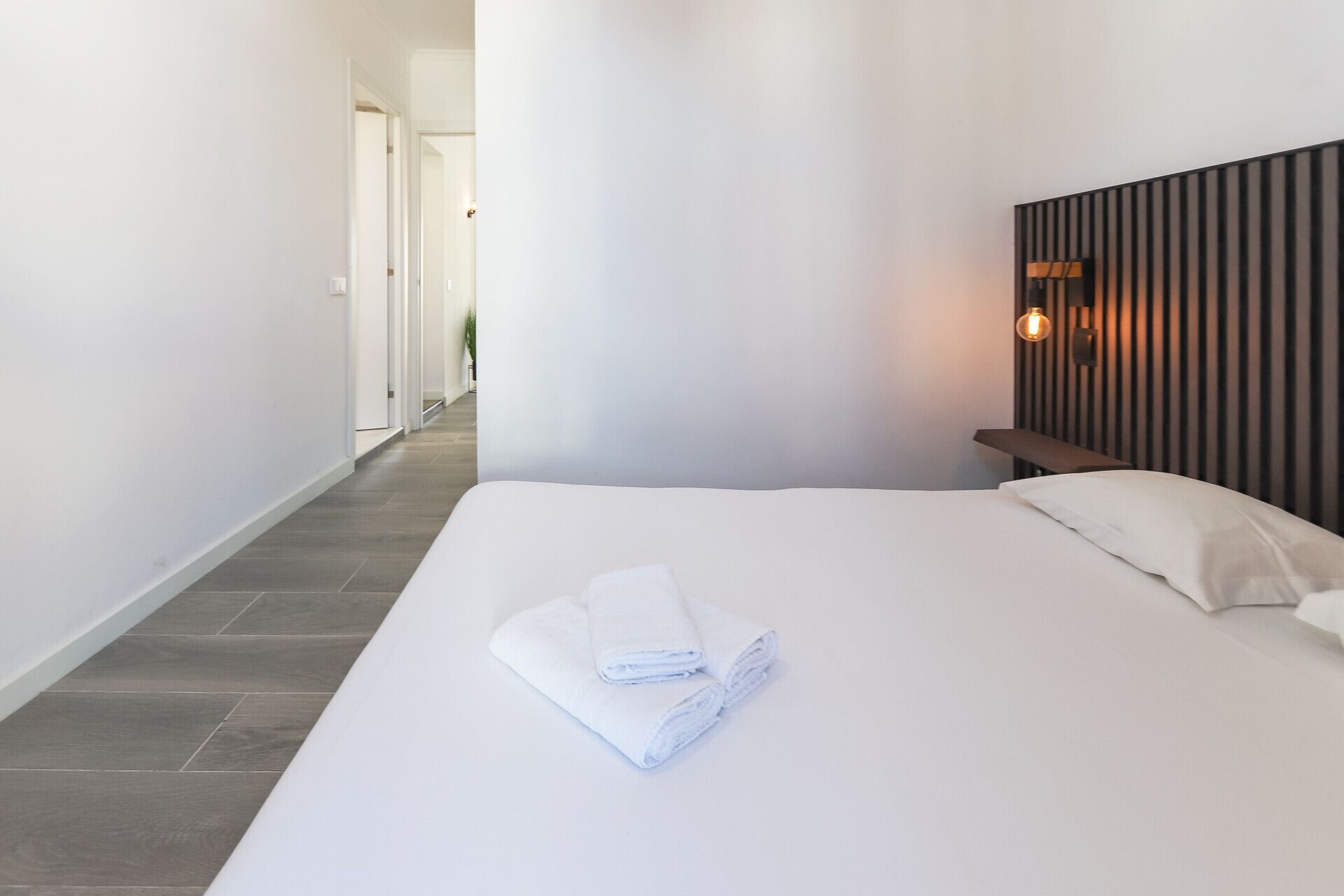 A hotel room with a large bed, a bedside table, a wall-mounted light, and a view of a hallway.
