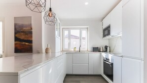Una cocina moderna con muebles blancos, horno empotrado y microondas. Hay una ventana que proporciona luz natural, y una lámpara colgante geométrica ilumina la encimera.