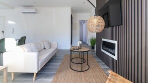Una sala de estar moderna con un sofá blanco, una mesa redonda negra, una lámpara de mimbre y un televisor montado en una pared oscura.