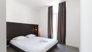 Habitación de hotel moderna con una cama grande, cabecera con líneas verticales, lámpara de noche y vistas al exterior a través de una ventana con cortinas.