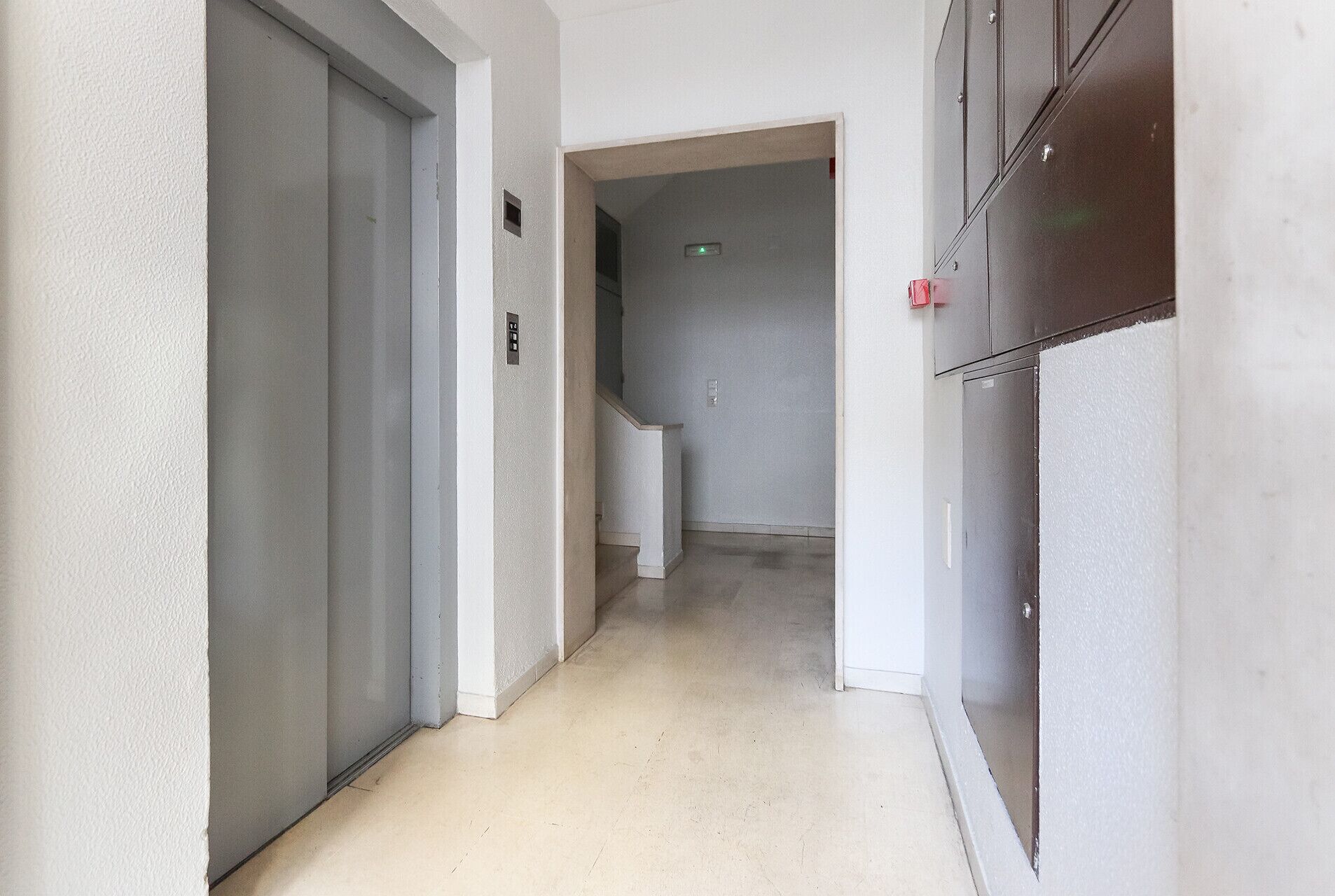 Un couloir étroit avec une porte blanche sur la gauche, une porte grise au centre et un casier métallique sur la droite.