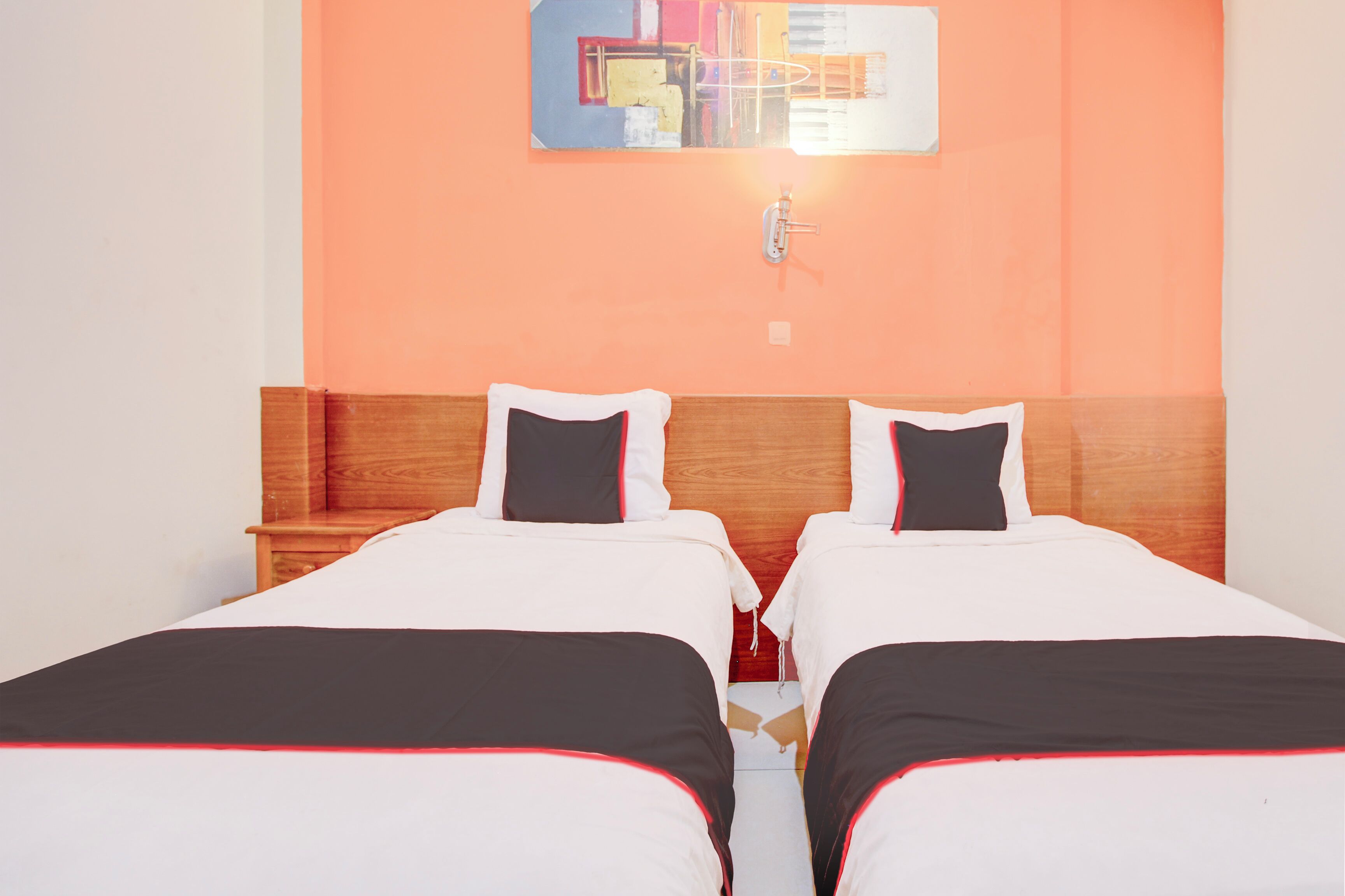 A hotel room with two single beds, wooden headboards, white bedding with red trim, and a colorful abstract painting on the wall.