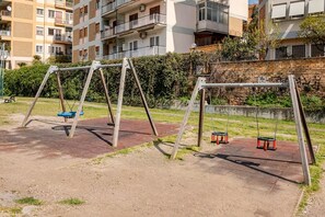 An outdoor playground with swings and a paved area, surrounded by apartment buildings.