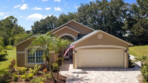 A two-story house with a garage, a front yard with various plants, and an American flag. - Private Pool Villa Near Major Theme Parks – Quiet Gated Golf Community (Haines City)