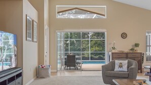 A spacious living room with a large TV, a grey armchair, a wooden coffee table, and a view of a swimming pool through glass doors. - Private Pool Villa Near Major Theme Parks – Quiet Gated Golf Community (Haines City)
