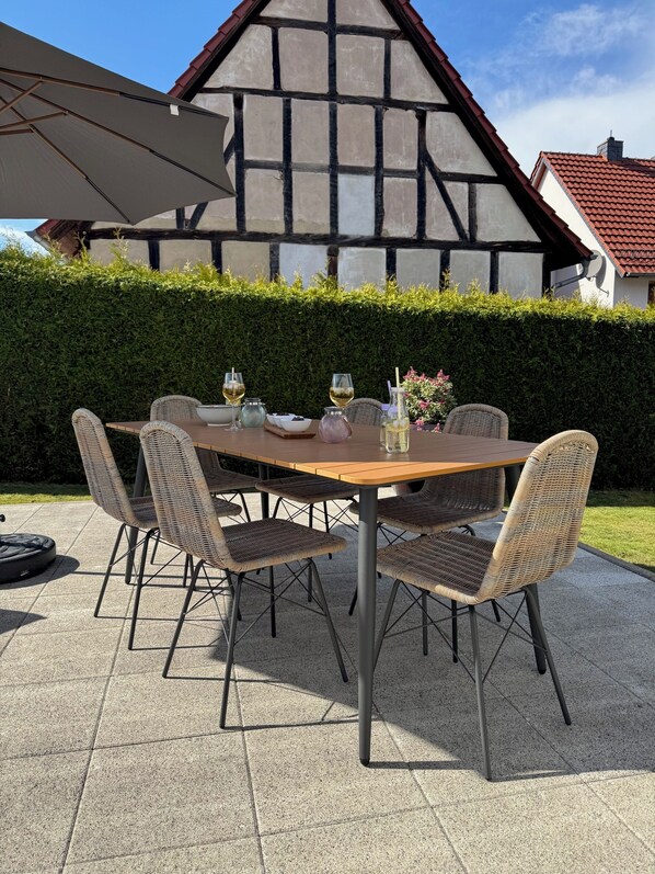 An outdoor dining area with a round table and wicker chairs, set against a backdrop of a half-timbered house and a neatly trimmed hedge.