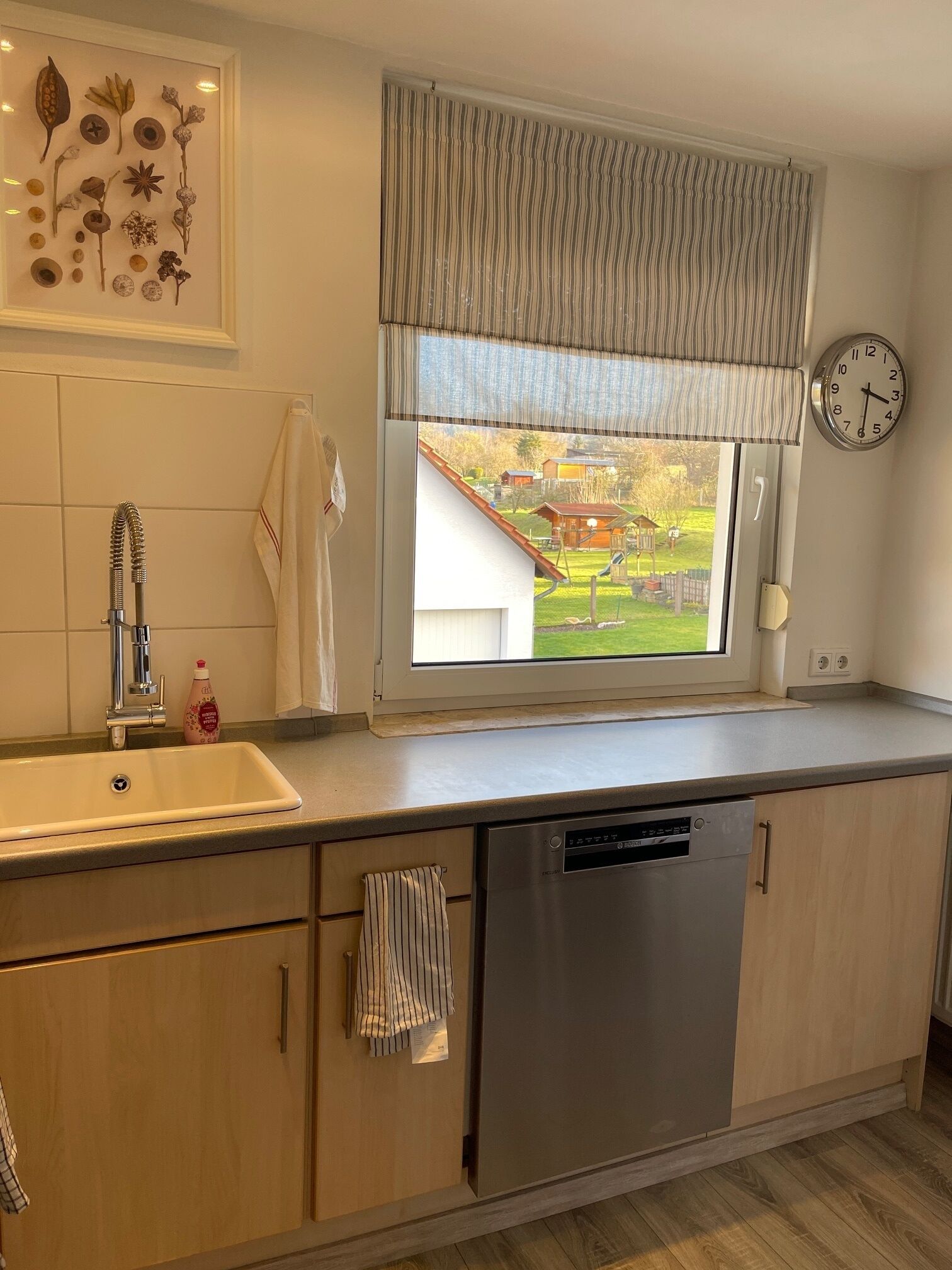 A modern kitchen with a built-in dishwasher, a window with a view of a house, and a wall-mounted clock.