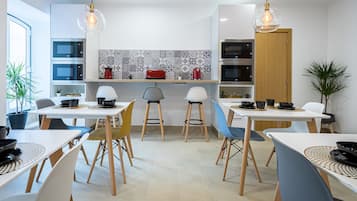 A modern kitchen with white cabinets, a tiled backsplash, and a wooden door. There are two dining tables with chairs, and a potted plant is visible.