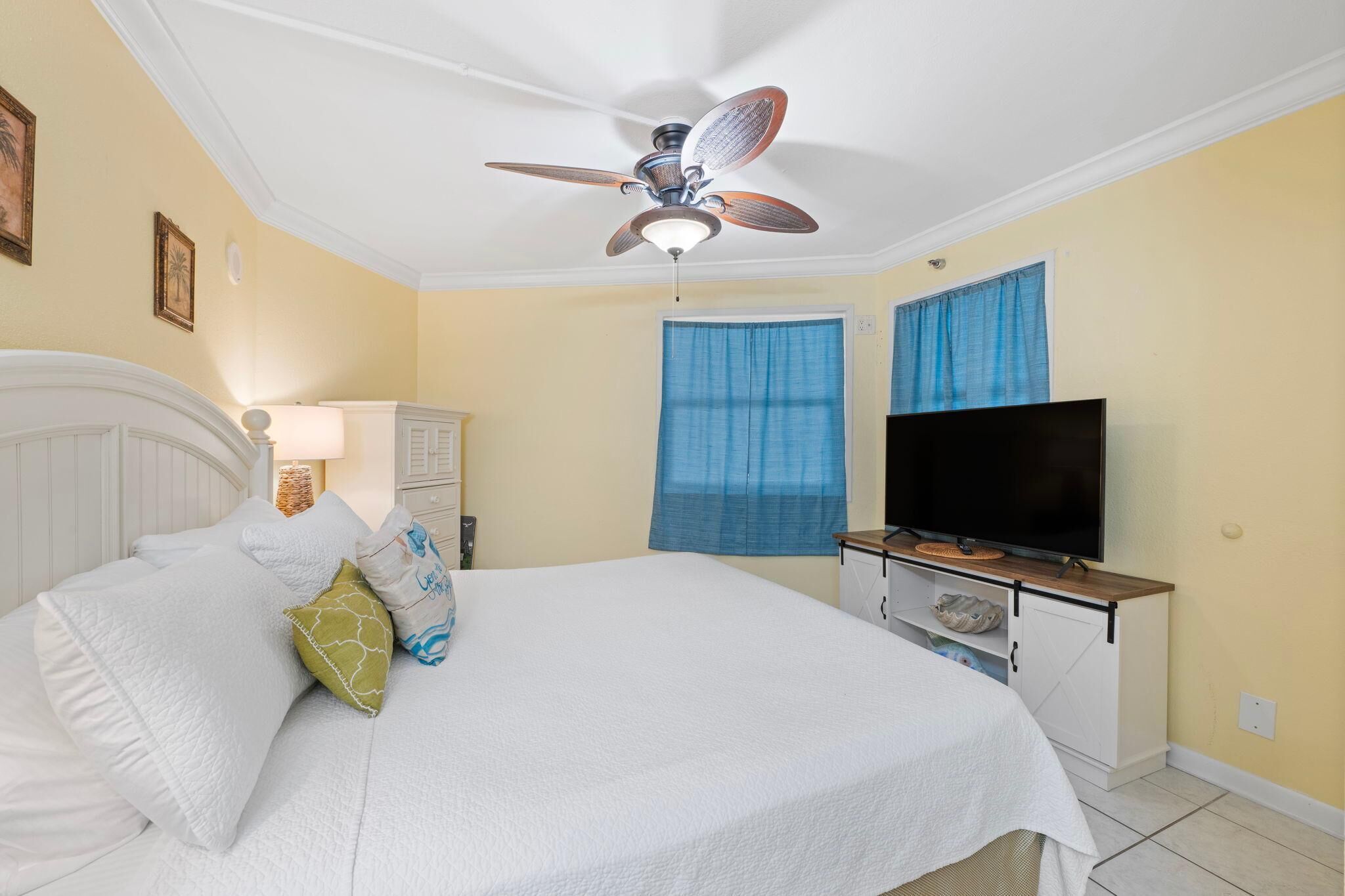 A bedroom with a bed, a ceiling fan, a TV, and a window with blue curtains.