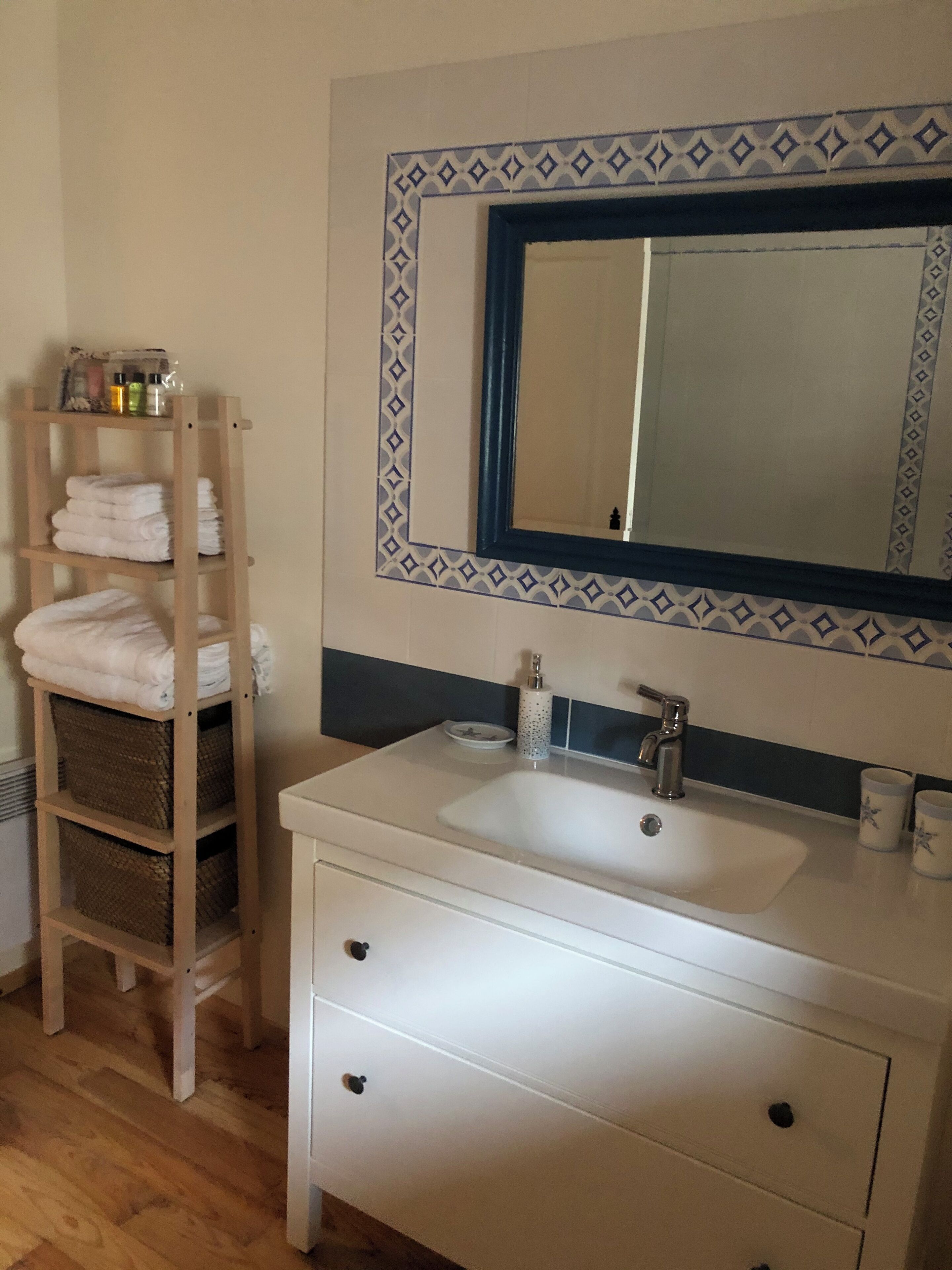 Een badkamer met een wit wastafelmeubel, een spiegel met decoratieve omlijsting, een handdoekrek met opgevouwen handdoeken en een plank met toiletartikelen.