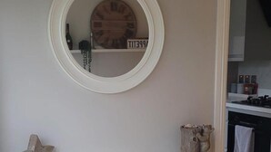 A round mirror with a wooden frame on a wall, a small shelf with a bottle and a decorative item, and a kitchen area with cabinets and appliances. - 3 Bedroom, Sleeps 6, Free Parking (Glasgow)