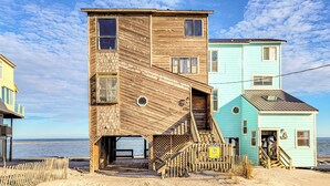 Et flerplans træhus med en trappe op til en terrasse, beliggende på en sandstrand med oceanen i baggrunden.