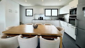 A modern kitchen with white cabinets, a wooden dining table, and a stainless steel refrigerator. - Villa d’architecte contemporaine - Vue mer 180° (Larmor-Plage)