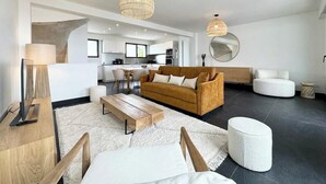 A modern living room with a sofa, coffee table, and ottoman, a kitchen in the background, and two hanging light fixtures. - Villa d’architecte contemporaine - Vue mer 180° (Larmor-Plage)