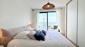 A modern bedroom with a large bed, a hanging light fixture, and a view of the sea through a large window. - Villa d’architecte contemporaine - Vue mer 180° (Larmor-Plage)