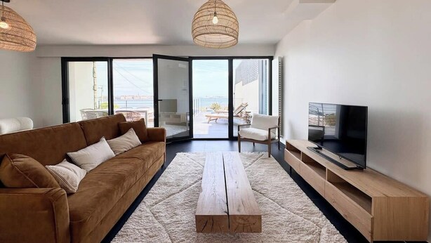A modern living room with a brown sofa, a wooden coffee table, a TV on a wooden stand, and a large window offering a view of the outdoors. - Villa d’architecte contemporaine - Vue mer 180° (Larmor-Plage)