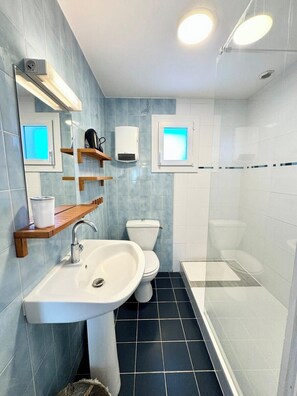 A bathroom with a sink, toilet, and shower. There are shelves above the sink and a window above the toilet. - Maison entièrement rénovée - À 2 pas des plages (Ploemeur)