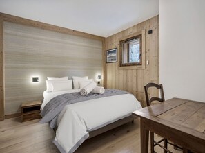 Ein Schlafzimmer mit Holz Kopfteil, einem Bett mit weißer Bettwäsche, einem kleinen Nachttisch, einem Holzstuhl und einem Fenster mit Blick auf Bäume.