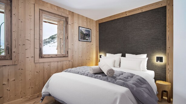 Ein Schlafzimmer mit einer Holzwand, einem großen Bett mit weißer Bettwäsche, einem kleinen Nachttisch und einem Fenster mit Blick auf eine verschneite Landschaft.