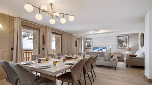 A modern dining area with a wooden table set for six, surrounded by upholstered chairs. A contemporary chandelier hangs above, and the room features a large window with a view of a snowy landscape.