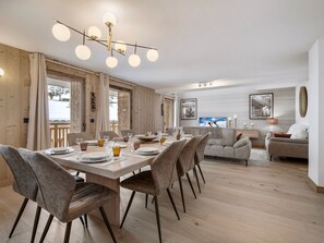 A modern dining area with a wooden table set for six, surrounded by upholstered chairs. A contemporary chandelier hangs above, and the room features a large window with a view of a snowy landscape. - Ski-in Ski-out Apartment for 10 with Wifi (Saint-Martin-de-Belleville)