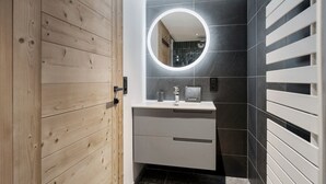 Ein modernes Badezimmer mit Holztür, rundem Spiegel, weißem Waschbecken und dunkel gefliesten Wänden.