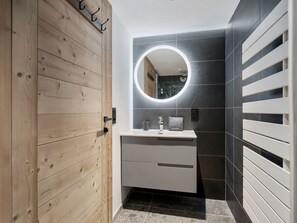Ein modernes Badezimmer mit Holztür, rundem Spiegel, weißem Waschbecken und dunkel gefliesten Wänden.