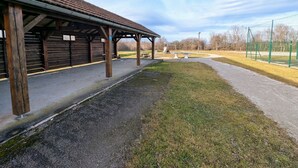 A covered outdoor area with benches and picnic tables, a fenced sports field, and a building with wooden beams and a tiled roof.