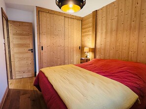 A bedroom with a wooden wardrobe, a bed with a red and yellow bedspread, and a nightstand with a lamp.