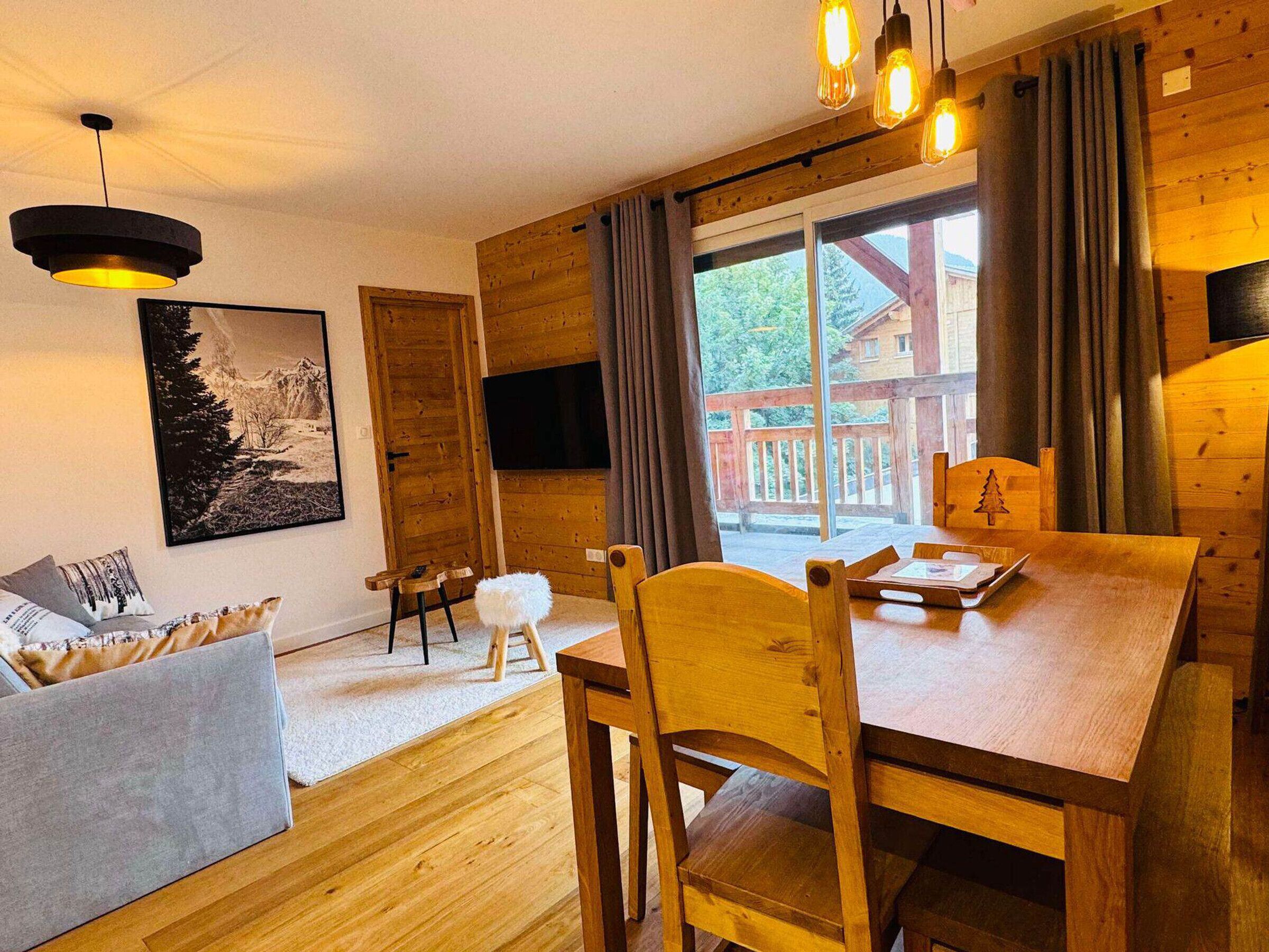 A cozy wooden interior with a dining table, chairs, a sofa, and a large window offering a view of a wooden deck outside.