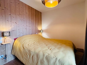 Quarto com cabeceira de madeira, cama com edredom bege, duas luminárias de mesa e uma tomada na parede.